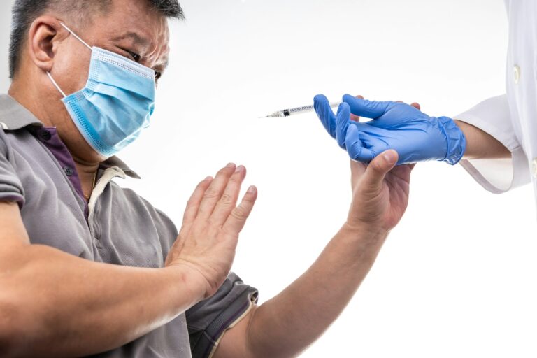 Fearful worried middle age man reacting to medical practitioner holding syringe loaded with Covid-19