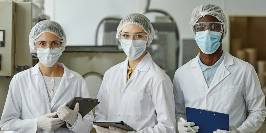 Three factory workers wearing lab coats and masks looking at camera in workshop