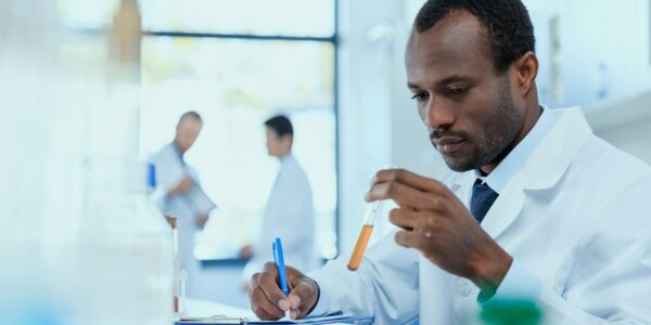 African american scientist in white coat holding and examining test tube with reagent, laboratory